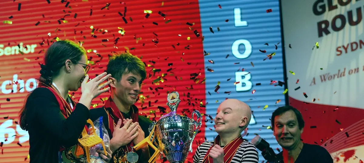 Three scholars celebrate with a trophy under falling confetti